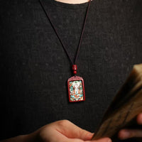 A person in a black shirt showcases Joyce’s Tibetjewelry Thangka Bodhisattva Manjushri Wealth Deity Necklace—Sacred Amulet for Prosperity & Spiritual Guidance—its vivid pendant on a black cord stands out as they hold blurred papers in the foreground.
